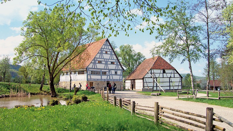 Bauernhaus und Scheune als Fachwerkbauten, links davor ein kleiner Teich. Auf dem Weg und vor dem Haus spazieren Besucher:innen im Schatten der Bäume