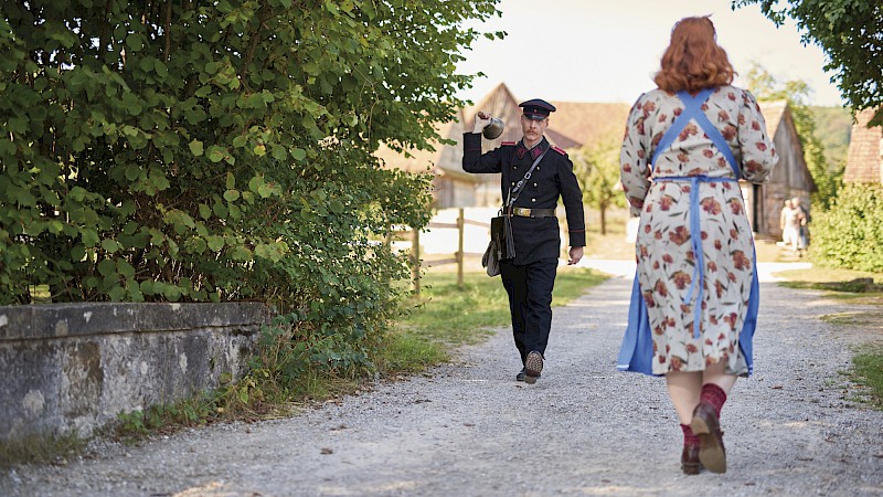 Nachgestellte historische Dorfszene der 1940er: Uniformierter Mann läutet im Laufen eine Glocke, eine Frau läuft ihm entgegen.