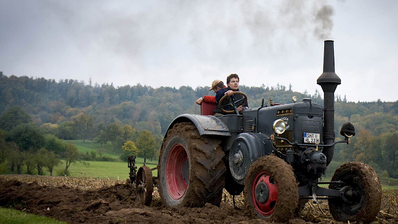 Ein historischer dunkelblauer Traktor wird von zwei Personen über den Acker gefahren