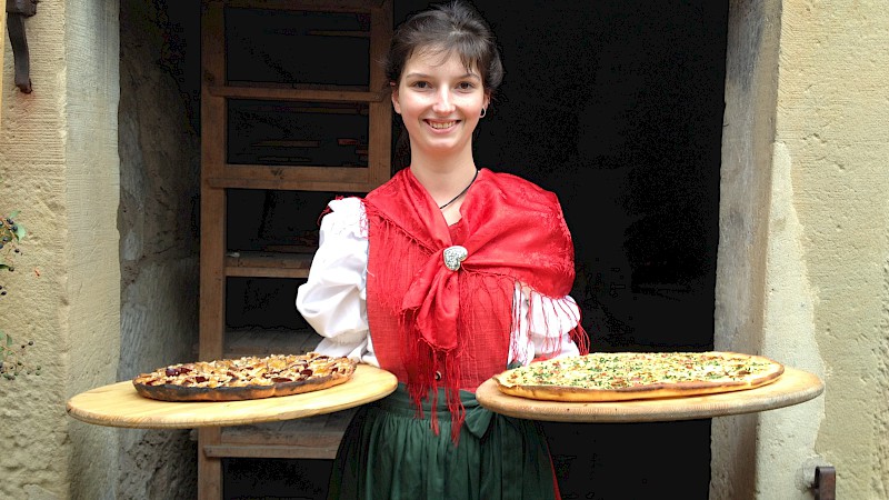 Junge Frau in einer Tracht rägt in je einer Hand ein großes Holztablett mit einem Blooz