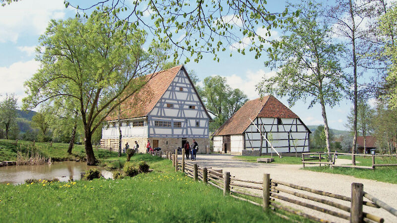 Bauernhaus und Scheune als Fachwerkbauten, links davor ein kleiner Teich. Auf dem Weg und vor dem Haus spazieren Besucher:innen im Schatten der Bäume.