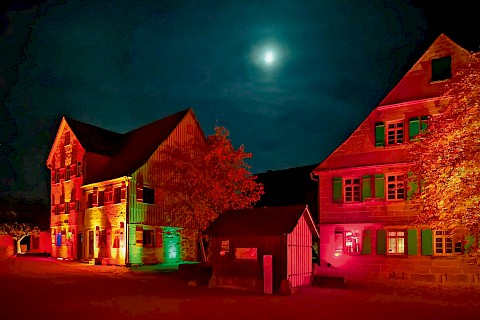 Zwei historische Steinhäuser und eine kleine Hütte werden bei Nacht mit rotem Licht angestrahlt