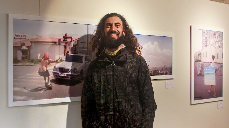 David Grigoryan steht vor seinen ausgestellten Fotografien und lacht in die Kamera. Das Bild neben ihm zeigt einen Skateboarder die Fahrbahn eines Autos schneidend