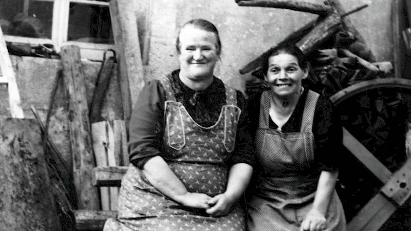 Historische Schwarz-Weiß-Fotografie zweier Frauen in Schürze, die in die Kamera lächeln. Gemeinsam sitzen sie auf einer Bank vor einem Haus