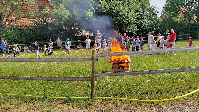Auf einer eingezäunten Wiese beobachten Besucher:innen eine sehr kleine brennende Holzhütte