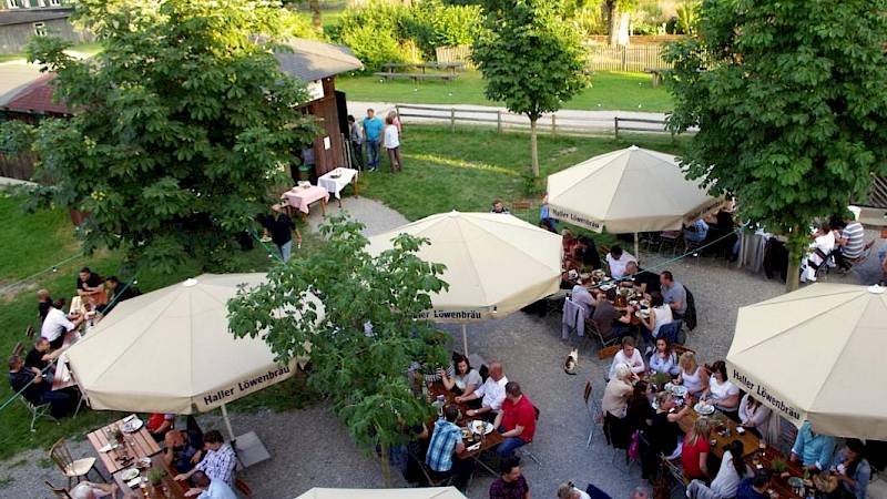 Biergarten von schräg oben, 5 beige Sonnenschirme, Schotterboden und vollbesetzte Biergarnituren.