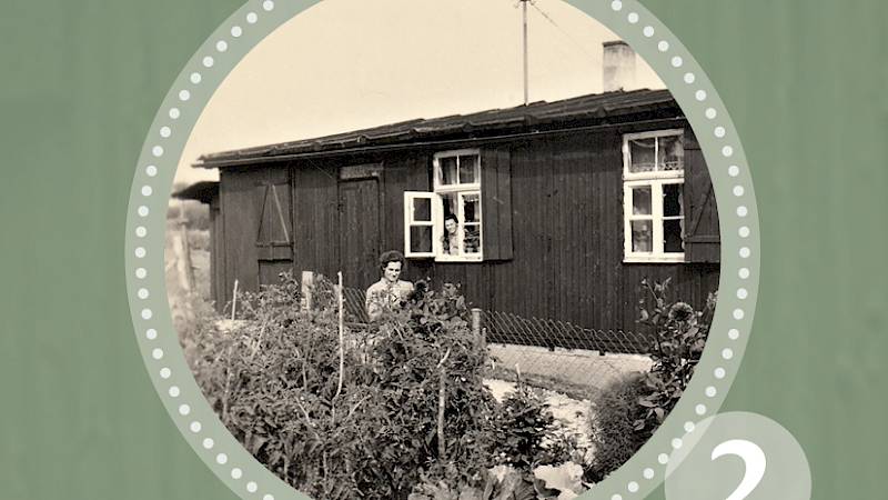 "Themenreihe Wohnbaracke aus Geschlachterbretzingen" 2. Historische Schwarz-Weiß-Fotografie einer alten Baracke als Wohnhaus umfunktioniert. Aus dem Fenster schaut eine Person heraus, vor dem Gebäude steht eine Frau am Zaun, welcher das Gelände eingrenzt