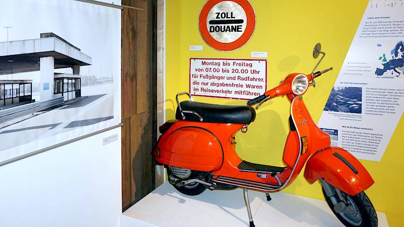 Ein historischer orangener Motorroller vor einer Ausstellungstafel. An der Tafel befinden sich neben alten Verkehrsschildern auch Fotografien und ein Ausstellungstext