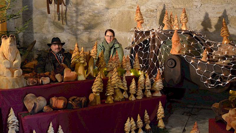 Zwei Verkäufer:innen hinter ihrem Vorweihnachtsmarktstand mit selbstgeschnitzer Holzdekoration
